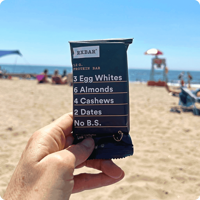 Hand holding an RXBAR protein bar at the beach — buy protein bars online from Misfits Market for on-the-go snacking