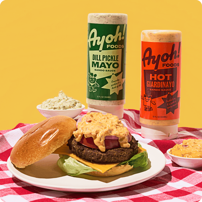 Ayoh Foods Dill Pickle Mayo and Hot Giardinayo sauces delivery beside a loaded cheeseburger with sauce drizzled on a picnic table.