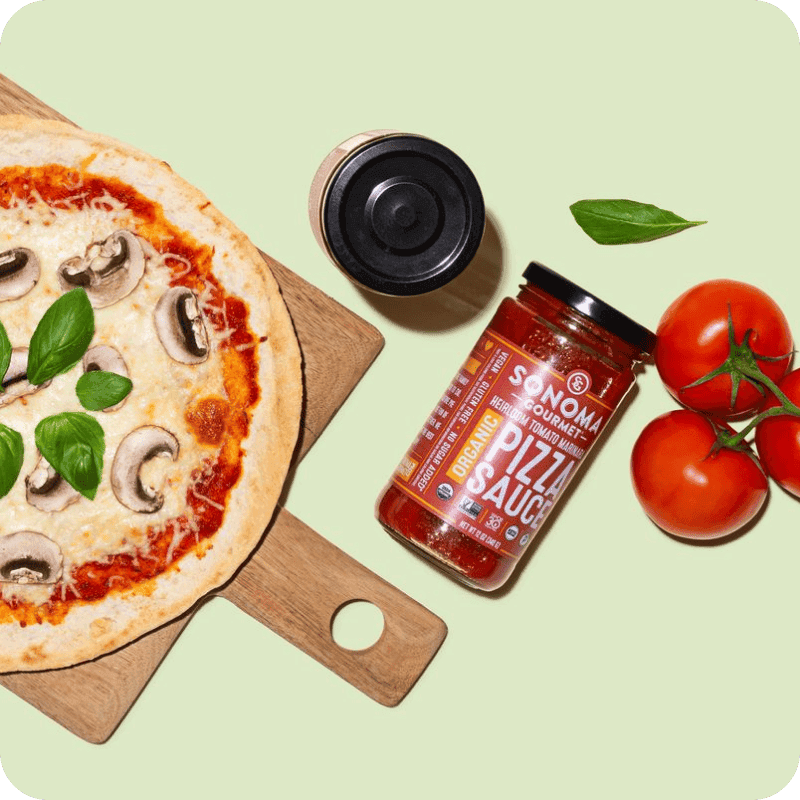 Sonoma Gourmet organic pizza sauce delivery jar beside a mushroom and basil pizza on a wood board with fresh vine tomatoes.
