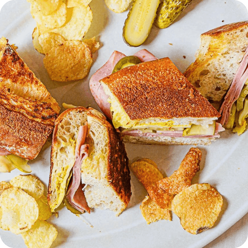Toasted sandwich with ham delivery, mustard, pickles, and melted cheese, sliced and served with chips and pickle spears."
