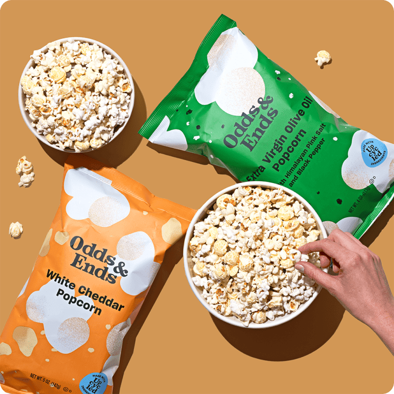 Odds & Ends White Cheddar and Extra Virgin Olive Oil gourmet popcorn bags with bowls of popcorn, available for popcorn delivery.