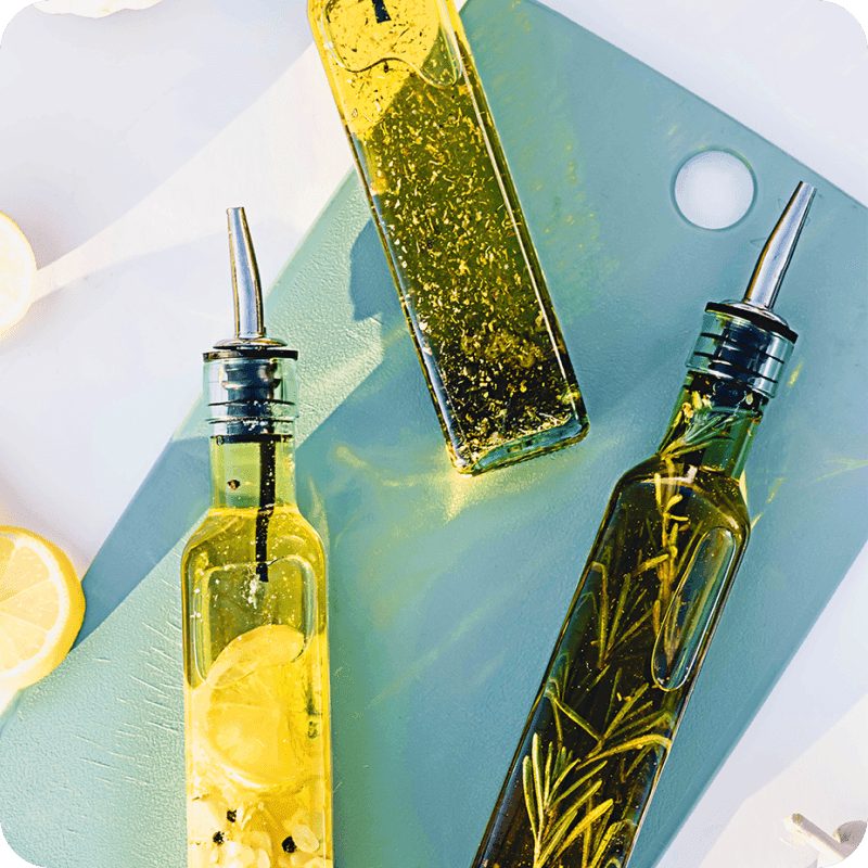 Glass bottles of herb and lemon infused olive oil with pour spouts on a cutting board, shop cooking oil delivery from Misfits Market.
