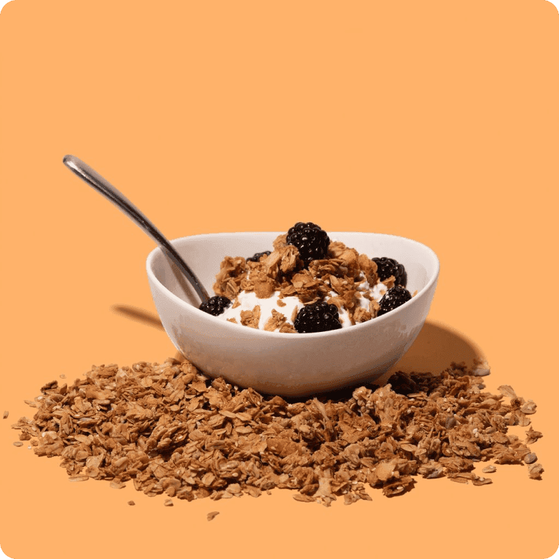 White bowl of yogurt delivery topped with granola and fresh blackberries, surrounded by scattered granola on an orange background.
