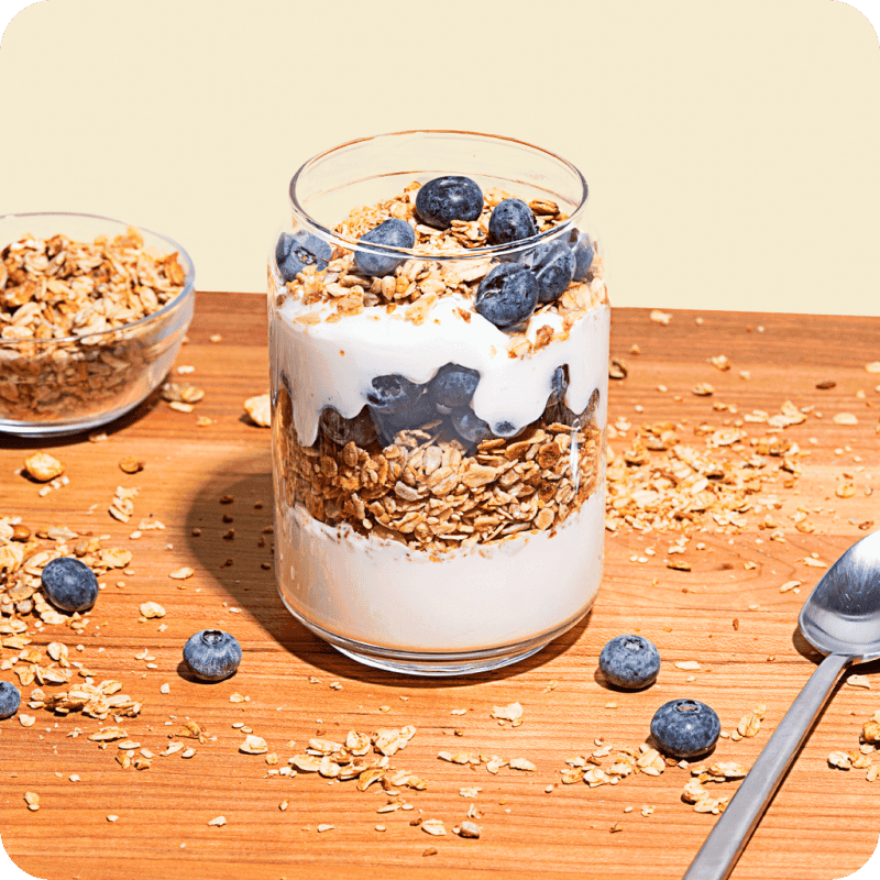 Greek yogurt delivery parfait layered with granola and fresh blueberries in a glass jar on a wooden surface.