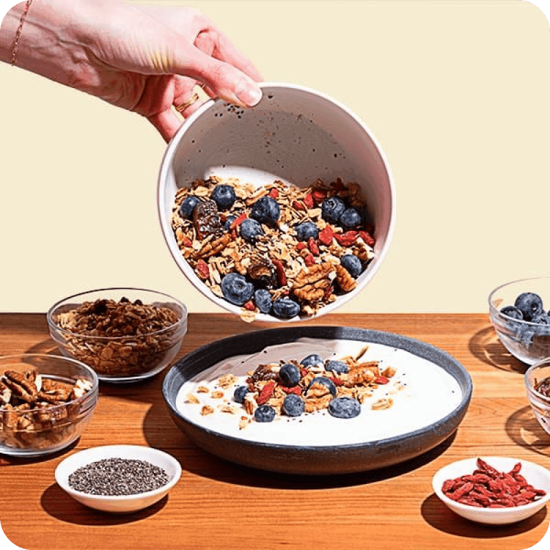 Pouring granola, blueberries, and goji berries over a yogurt delivery bowl topped with chia seeds, pecans, and mixed toppings.