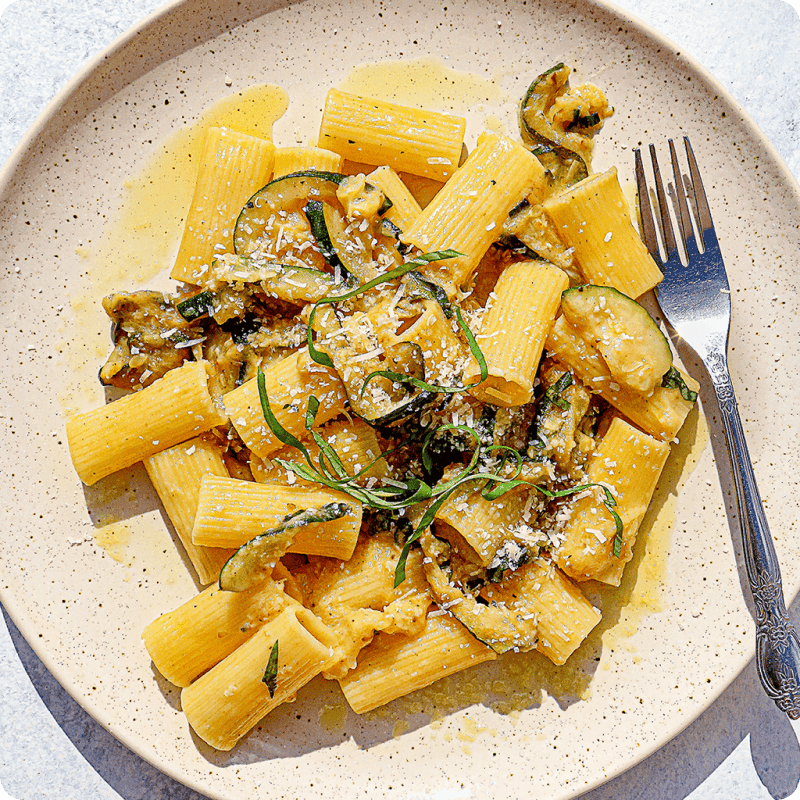 Rigatoni pasta delivery dish tossed with sautéed zucchini, fresh basil, and grated parmesan on a speckled ceramic plate.