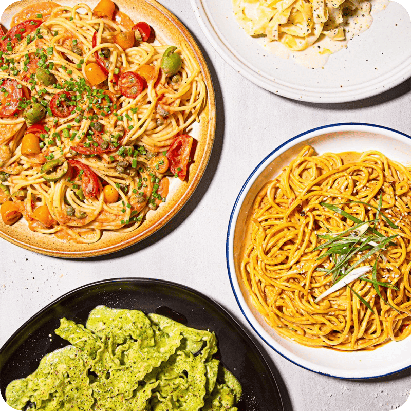 Four pasta delivery dishes including tomato spaghetti, creamy white tagliatelle, pesto pasta, and golden herb noodles overhead view.