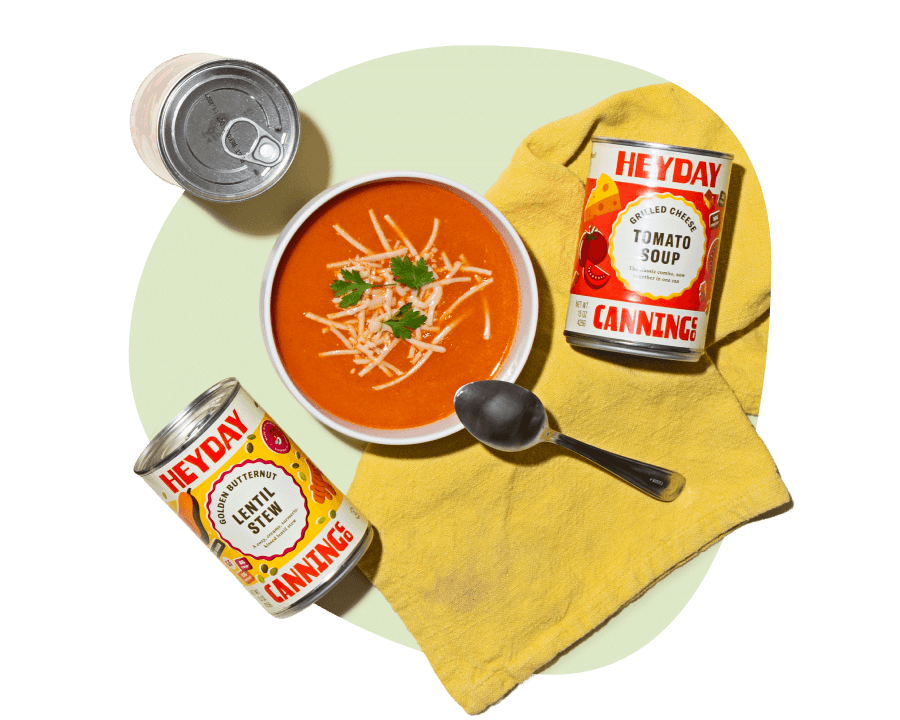 Heyday Canning Co. tomato soup with shredded cheese in a bowl, alongside cans of tomato soup and lentil stew from a gourmet soup delivery service.