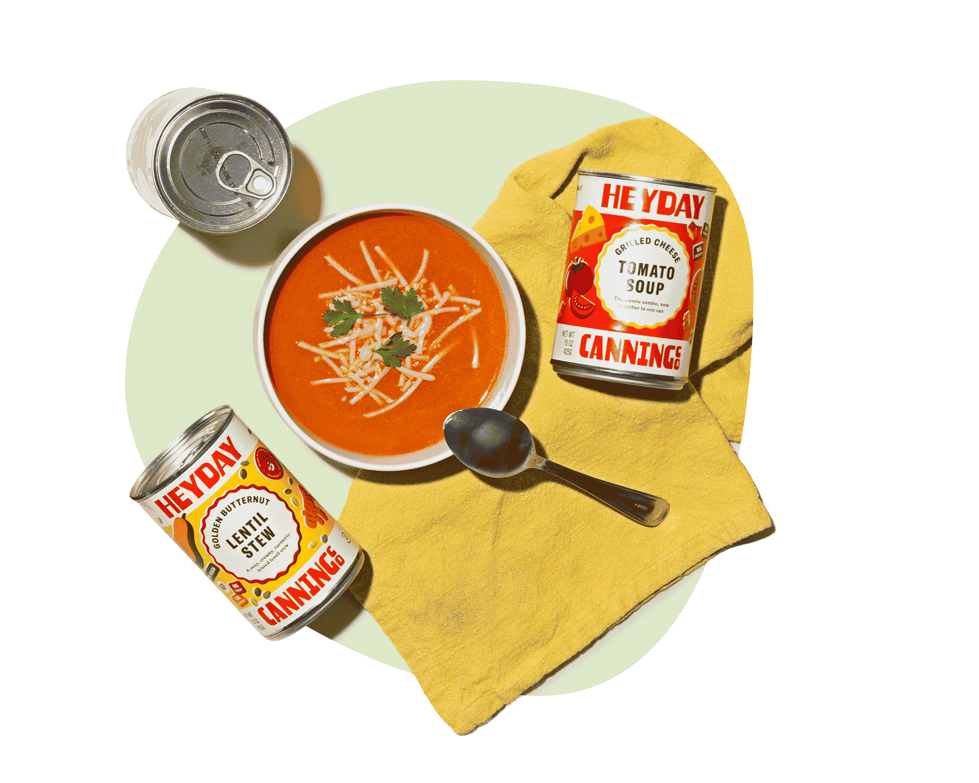 Heyday Canning Co. tomato soup with shredded cheese in a bowl, alongside cans of tomato soup and lentil stew from a gourmet soup delivery service.